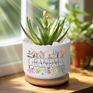 May include: White ceramic planter with a floral design and the word "Grandma" with three names below it. The planter has a bamboo base and is filled with a green succulent plant.