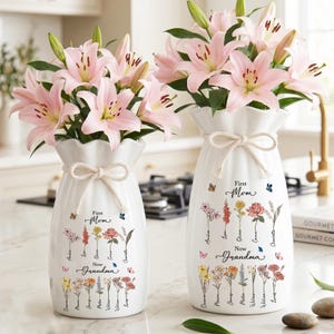 May include: Two white ceramic vases with pink lilies, tied with a bow. The vases are decorated with floral illustrations and the text "First Mom Now Grandma". Butterflies are also depicted on the vases.