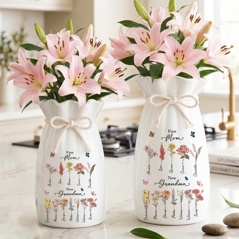 May include: Two white ceramic vases with pink lilies, tied with a bow. The vases are decorated with floral illustrations and the text "First Mom Now Grandma". Butterflies are also depicted on the vases.