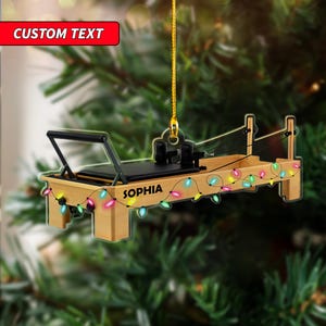 May include: A wooden Pilates reformer Christmas ornament with the name "SOPHIA" and colorful string lights. The ornament has a black seat and a gold hanging string. The top left corner has a red banner that says "CUSTOM TEXT".