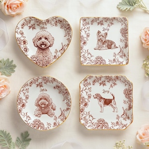 May include: Four decorative ceramic plates with dog illustrations. The plates are in various shapes: heart, square, round, and a scalloped square. Each plate features a different dog breed, surrounded by floral patterns and a gold rim. The dogs' names are printed above the illustrations.