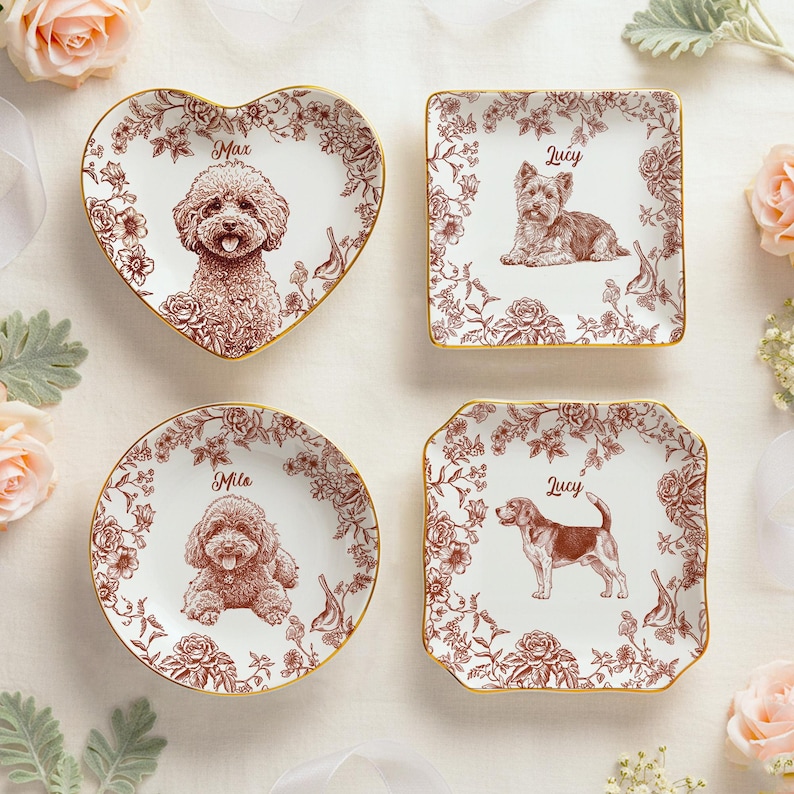 May include: Four decorative ceramic plates with dog illustrations. The plates are in various shapes: heart, square, round, and a scalloped square. Each plate features a different dog breed, surrounded by floral patterns and a gold rim. The dogs' names are printed above the illustrations.