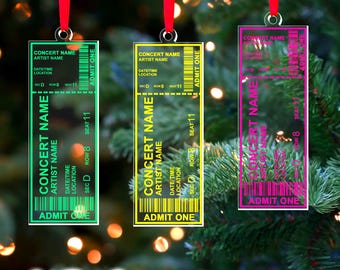 May include: Three concert ticket-shaped Christmas ornaments with red ribbons. The ornaments are in green, yellow, and pink, each with a different color border and text. The text includes "CONCERT NAME", "ARTIST NAME", and "ADMIT ONE".