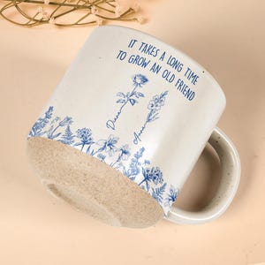 May include: Cream-colored ceramic mug with a speckled handle and a textured, unglazed base. The mug features blue floral illustrations and the text "IT TAKES A LONG TIME TO GROW AN OLD FRIEND" with the names "Diana" and "Anna".