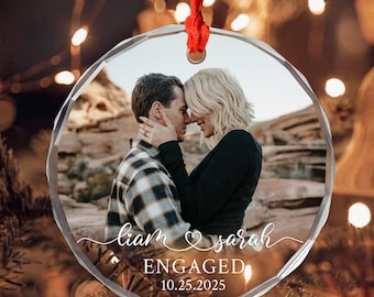 May include: A clear, faceted glass ornament with a red ribbon. The ornament features a photo of a couple, with the words "Liam & Sarah" and "ENGAGED 10.25.2025" in white script. The couple is embracing, with a natural landscape in the background.