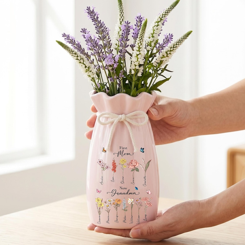May include: A pink ceramic vase with a ruffled top and a tied bow detail, filled with purple and white flowers. The vase is decorated with floral illustrations and the text "First Mom Now Grandma".