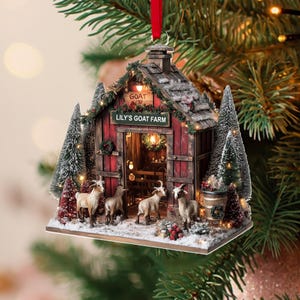 May include: A Christmas ornament featuring a red barn with the text "LILY'S GOAT FARM". The barn has a wooden roof, a small sign that says "GOAT", and is surrounded by miniature evergreen trees and goats. The ornament is hanging from a red ribbon.