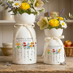 May include: Two white ceramic vases with floral arrangements. The vases have a scalloped top and are tied with a cream-coloured ribbon. One vase reads "Grandma's Garde" and the other reads "Nana's Garden" with names below.