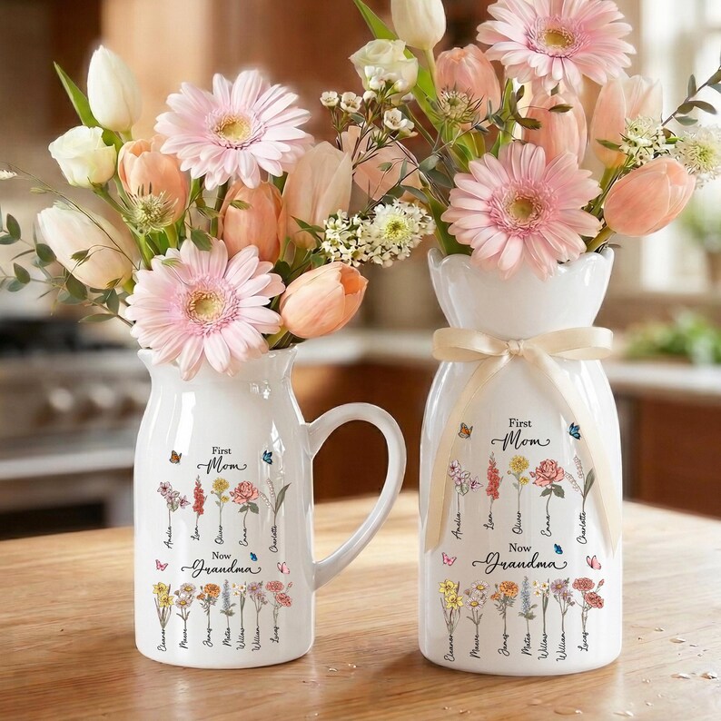 May include: Two white ceramic vases with floral arrangements. The vases are decorated with the words "First Mom, Now Grandma" and floral illustrations. One vase has a handle. The flowers are pink, peach, and white.