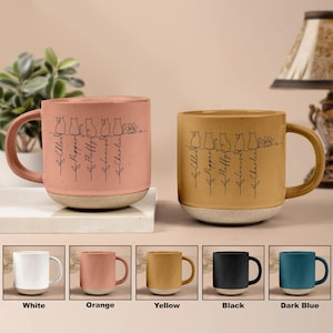 May include: Two ceramic mugs with a cat design and names. One is pink with a white base, the other is yellow with a white base. The mugs have a handle and are decorated with a line drawing of cats and names. Below are color options: white, orange, yellow, black, and dark blue.