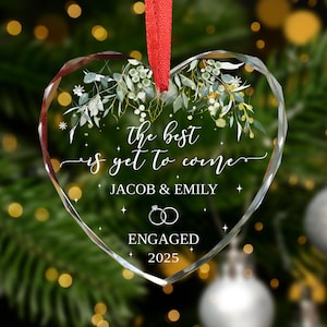 May include: A heart-shaped clear glass ornament with a red ribbon. The ornament has the text "the best is yet to come" with the names "JACOB & EMILY", the word "ENGAGED", and the year "2025". The ornament is decorated with green and gold floral accents.