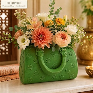 Peut inclure: Un vase vert en forme de sac à main imprimé en 3D en PLA avec un motif floral en relief et deux anses, rempli d'un arrangement vibrant de fleurs pêche, blanches et jaunes, incluant des dahlias, des roses et du feuillage d'eucalyptus.