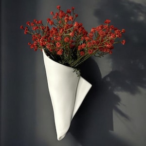 May include: A white ceramic cone-shaped vase with a red and white flower arrangement. The vase is mounted on a dark grey wall.
