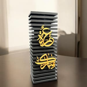 May include: A grey, stacked, rectangular decorative stand with two gold Arabic calligraphy words on it. The stand is on a brown surface.
