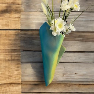Peut inclure: Un bouquet de fleurs blanches dans un vase conique bleu et vert. Le vase est fait d'un matériau texturé et présente un dégradé de couleur du bleu au vert. Le vase est suspendu à un mur en bois.