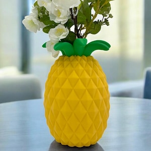 May include: A yellow pineapple-shaped vase with a green top holds white flowers and green leaves. The vase has a textured surface and sits on a gray table. The background is blurred, with a window and chairs visible.