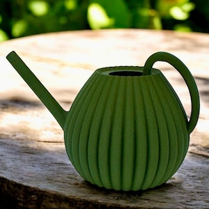 Könnte beinhalten: Eine grüne Gießkanne mit geripptem Design und einem gebogenen Griff. Die Gießkanne steht auf einer Holzoberfläche.