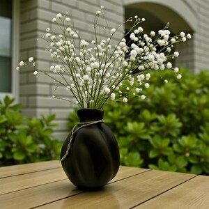 May include: A matte black vase with a rounded, ribbed body and a narrow neck, featuring a thin, light-coloured twine wrapped around the top. The vase is filled with a large, airy bouquet of small, white baby's breath flowers on thin green stems.