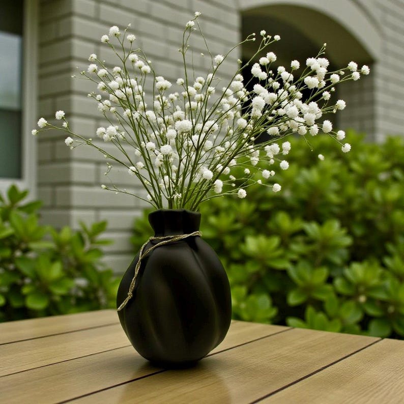 May include: A matte black vase with a rounded, ribbed body and a narrow neck, featuring a thin, light-coloured twine wrapped around the top. The vase is filled with a large, airy bouquet of small, white baby's breath flowers on thin green stems.