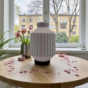 Puede incluir: Una lámpara de cerámica blanca y acanalada con base negra sobre una mesa de madera. Pétalos de flores rosas esparcidos alrededor de la lámpara. Un pequeño jarrón con flores rosas a la izquierda. La lámpara mide unos 25 cm de altura.