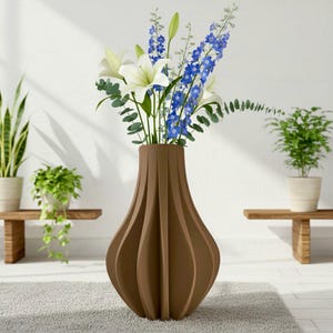 Peut inclure: Un vase marron nervuré rempli de lys blancs, de delphiniums bleus et d'eucalyptus verts. Le vase présente un design incurvé unique et repose sur un tapis gris. Des plantes en pot sont visibles en arrière-plan, ajoutant à l'esthétique naturelle.