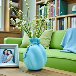 May include: A light blue, rounded ceramic vase with a cinched neck and a thin, metallic gold cord wrapped around the top. The vase contains a bouquet of tall, slender green stems topped with small, delicate white flowers.