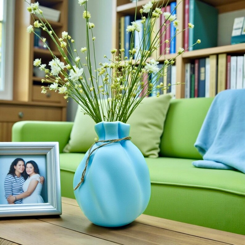 May include: A light blue, rounded ceramic vase with a cinched neck and a thin, metallic gold cord wrapped around the top. The vase contains a bouquet of tall, slender green stems topped with small, delicate white flowers.