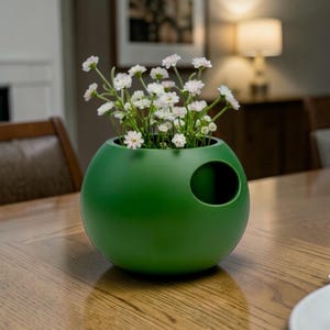 Puede incluir: Un jarr&oacute;n verde mate de forma esf&eacute;rica, con una abertura lateral, que contiene flores blancas y rosas. Colocado sobre una mesa de madera, aporta un toque moderno a la decoraci&oacute;n del hogar. Dise&ntilde;o &uacute;nico.