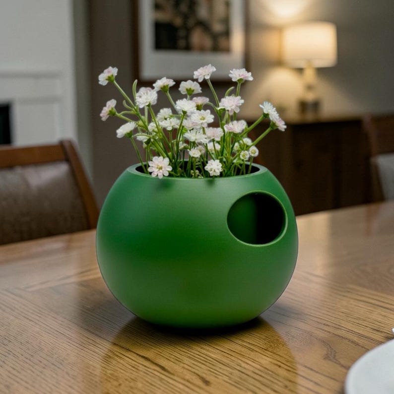 Puede incluir: Un jarr&oacute;n verde mate de forma esf&eacute;rica, con una abertura lateral, que contiene flores blancas y rosas. Colocado sobre una mesa de madera, aporta un toque moderno a la decoraci&oacute;n del hogar. Dise&ntilde;o &uacute;nico.