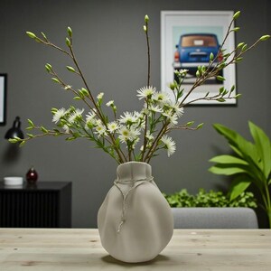 May include: A matte light grey ceramic vase with an organic, rounded, and pinched shape. A thin, light-coloured string is tied around the neck of the vase. Inside are several branches with green buds and clusters of small, white, star-shaped flowers.