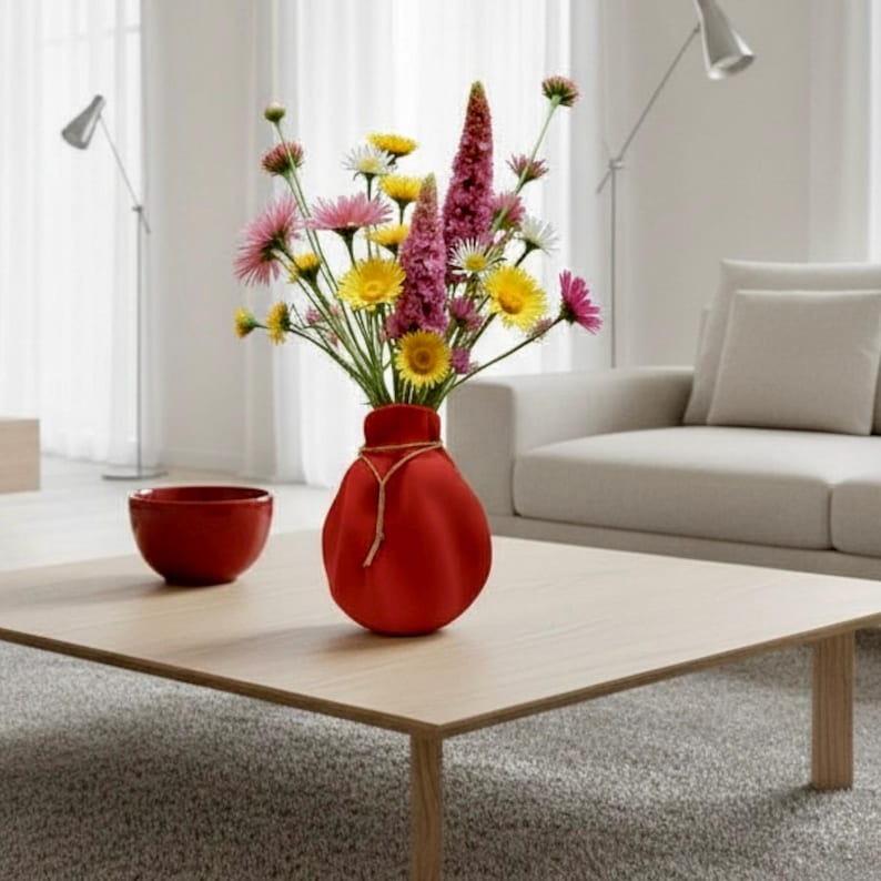 May include: A vibrant bouquet of mixed wildflowers featuring pink, yellow, and white petals arranged in a rounded, matte red vase. The vase is cinched at the neck with a thin, light-coloured cord tied in a simple knot, creating a soft, organic shape.
