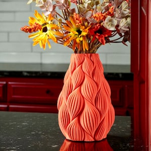 May include: A coral-coloured vase with a textured, leaf-like pattern, filled with an arrangement of orange, yellow, and red flowers. The vase is the focal point, set against a dark countertop and a red cabinet.
