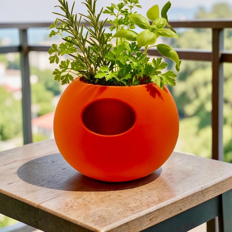 Puede incluir: Una maceta esf&eacute;rica naranja brillante con una abertura ovalada, llena de hierbas frescas. La maceta est&aacute; sobre una mesa de madera, con un balc&oacute;n y vegetaci&oacute;n al fondo. Las hierbas incluyen romero, perejil y albahaca.