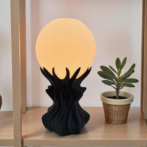 May include: A decorative table lamp with a large, spherical, glowing light. The light is supported by a black, stylized base with a textured, organic design. A small potted plant sits nearby on a wooden shelf.