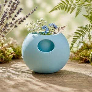 Puede incluir: Un jarr&oacute;n esf&eacute;rico azul claro con una abertura circular en el lateral. El jarr&oacute;n contiene un peque&ntilde;o arreglo de flores blancas y azules. El fondo presenta vegetaci&oacute;n y una superficie de madera.