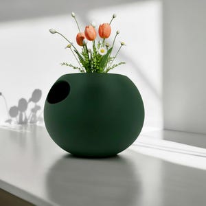 Puede incluir: Un jarr&oacute;n esf&eacute;rico verde oscuro con una abertura circular en el lateral. El jarr&oacute;n contiene un peque&ntilde;o arreglo de tulipanes naranjas, margaritas blancas y vegetaci&oacute;n. El jarr&oacute;n est&aacute; sobre una superficie blanca.