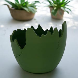 Puede incluir: Un macetero de cerámica verde con un borde agrietado e irregular. El macetero tiene forma de huevo y un acabado liso y mate.