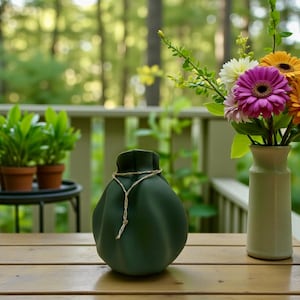 May include: A matte dark green ceramic vase with a rounded, pleated body and a narrow neck. A thin, light-coloured braided cord is tied around the neck of the vase, with the ends hanging down the front. The vase sits on a wooden surface.