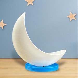 Op de afbeelding: Een wit keramisch nachtlampje in de vorm van een halve maan met een blauwe basis. Het nachtlampje staat op een houten oppervlak.