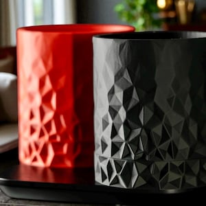 May include: Two cylindrical planters with geometric patterns. One is a glossy red, the other is matte black. Both are on a black tray, with a blurred background. The planters have a textured, faceted surface.