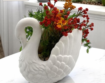 Puede incluir: Una maceta blanca con forma de cisne impresa en 3D, hecha de PLA y no de cerámica, que contiene un ramo de flores artificiales rojas y naranjas con follaje verde. El cisne presenta plumas moldeadas detalladas y un cuello curvado.