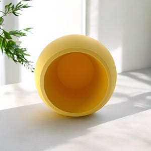 Puede incluir: Una maceta esf&eacute;rica amarilla con una amplia abertura. El interior es de un tono amarillo m&aacute;s oscuro. La maceta est&aacute; sobre una superficie blanca, con follaje verde en el fondo. Es un objeto decorativo.