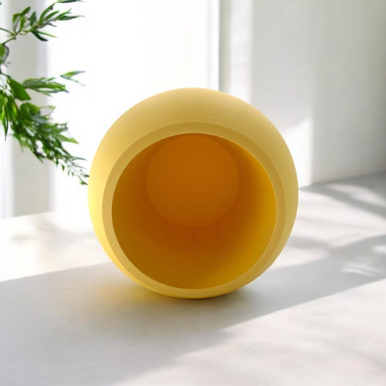 Puede incluir: Una maceta esf&eacute;rica amarilla con una amplia abertura. El interior es de un tono amarillo m&aacute;s oscuro. La maceta est&aacute; sobre una superficie blanca, con follaje verde en el fondo. Es un objeto decorativo.