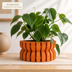 May include: A 3D printed orange planter with a ribbed, cylindrical design containing a green heart-leaf philodendron plant. The planter features a label that reads 3D PRINTED Premium PLA Not Ceramic. The pot has a distinct two-tier structure.