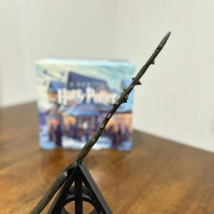 May include: A black wand with a dark wood finish and a pointed tip is displayed on a black triangular stand with the Deathly Hallows symbol. The wand is leaning against the stand, with the tip pointing upwards. The stand is on a wooden surface.