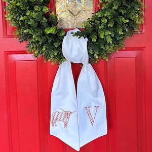 May include: A white fabric door wreath with a red embroidered longhorn steer and the letter "V" hanging on a red door with a green wreath.