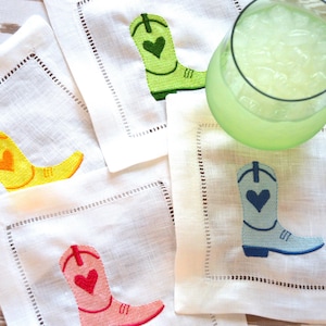 May include: Four white linen napkins with embroidered cowboy boots in different colors: green, yellow, pink, and blue. Each boot has a small heart embroidered on the top.