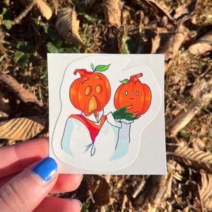 May include: A sticker featuring a cartoon pumpkin with a surprised expression holding another smaller pumpkin with a sad expression. The pumpkins are orange with green stems and leaves.