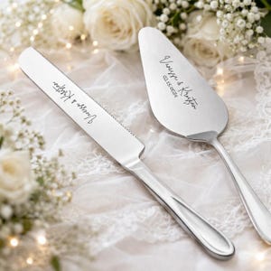 May include: A silver cake knife and server set. The knife has a serrated edge and the server is triangular. Both are engraved with the names "Vanessa & Kristian" and the date "02.15.2024". The set rests on a white lace surface.