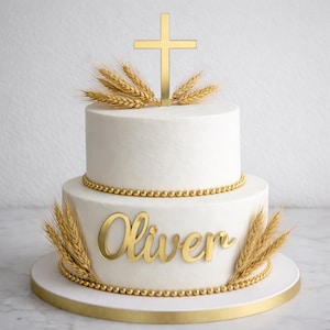 May include: A two-tiered white cake decorated with gold accents. A gold cross and wheat stalks adorn the top. The name "Oliver" is displayed in gold script on the bottom tier. The cake is on a white and gold cake stand.